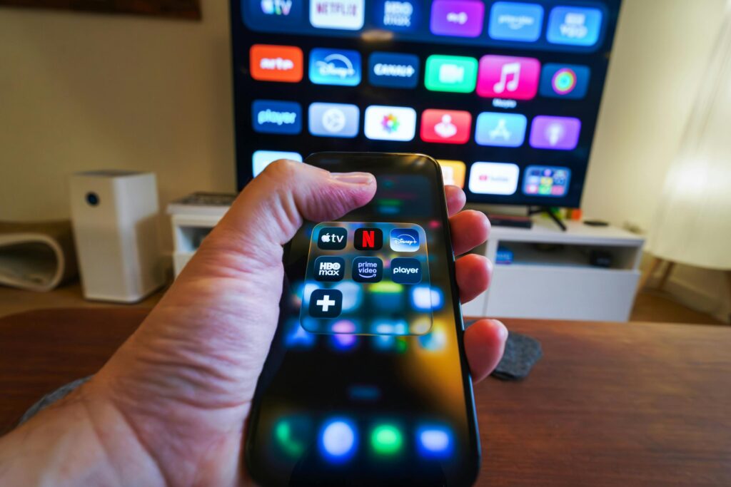 Close-up of a hand holding a phone displaying streaming apps in front of a TV with multiple app icons.